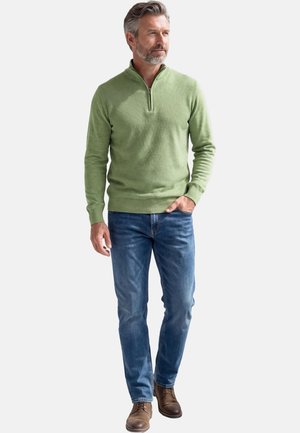 Mężczyzna o siwych włosach i brodzie, ubrany w jasnooliwkowy sweter z suwakiem do połowy wysokości, niebieskie dżinsy i brązowe skórzane buty, stojący z jedną ręką w kieszeni.