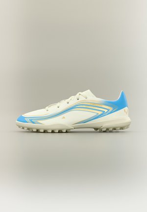 Bota de fútbol blanca con detalles en azul y dorado, cordones y tachones de goma en la suela, mostrada sobre un fondo neutro.