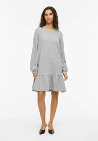 Robe grise à manches longues avec un col rond, un ourlet volanté froncé et une texture douce. Associée à des chaussures noires à bouts pointus.