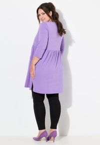 Lila tunika med insydd midja, trekvarts ärmar och sidofickor, som bärs med svarta leggings och lila högklackade skor.