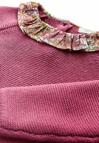 Pull en tricot rose avec un col en tissu à volants et motif floral dans des tons de rose, blanc, jaune et vert.