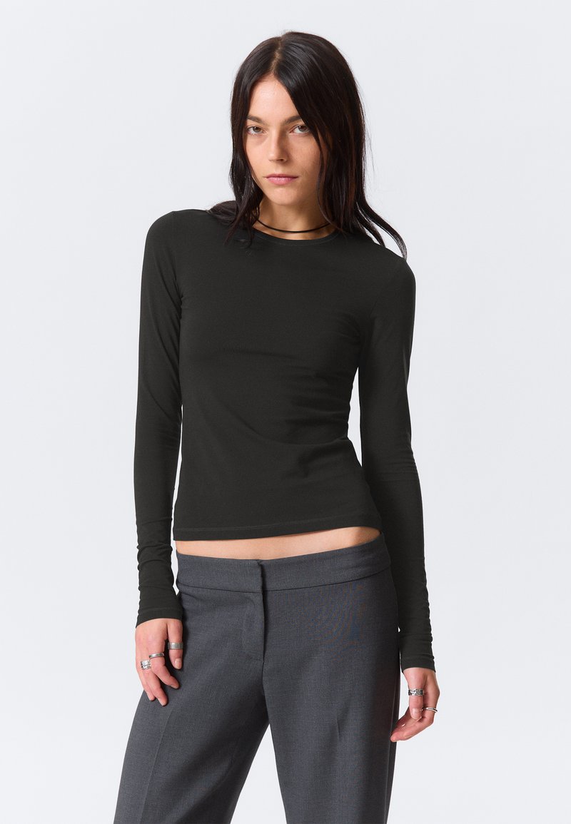 Młoda kobieta z długimi ciemnymi włosami, ubrana w czarną dopasowaną bluzkę z długim rękawem oraz szare spodnie z wysokim stanem, stoi na jednolitym tle.