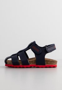 Lasten tummansininen sandaali, jossa on kolme etuhetiä, mokkanahkainen korkkijalkapohja, punainen teksturoitu pohja ja säädettävä takahihna tarranauhalla.