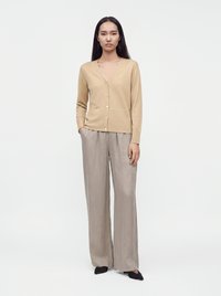 Femme aux longs cheveux foncés porte un cardigan beige boutonné, un pantalon ample gris clair et des chaussures noires pointues, debout devant un fond blanc.