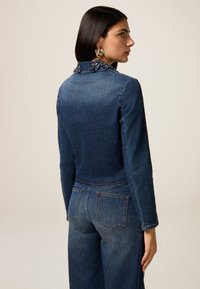 Veste en denim bleu foncé avec une taille cintrée, des manches longues et un col orné. Associée à un jean large assorti avec des poches arrière.