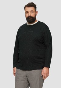 Schwarzes Langarmshirt mit gesprenkeltem Muster, mit Rundhalsausschnitt und einem dezenten Textdesign auf der Vorderseite. Getragen mit grauen Hosen.