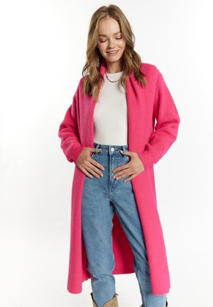 Lungo cardigan rosa in tessuto ribbed, aperto davanti, indossato sopra un top bianco e jeans blu a vita alta. Il modello posa con le mani sui fianchi, sorridendo.