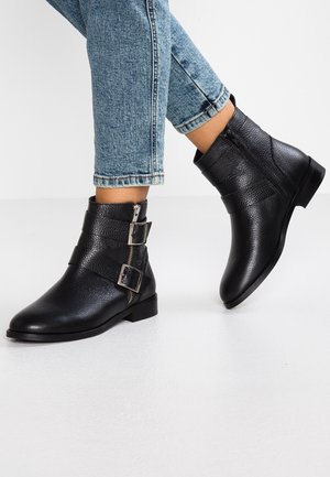 Bottines en cuir noir avec une finition texturée, dotées de deux accents de boucles en argent et d'une fermeture éclair sur le côté. Portées avec un jean bleu retroussé.