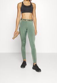 Czarny sportowy biustonosz, zielone legginsy z wysokim stanem z logo HKMY oraz czarne buty sportowe. Modelka trzyma lekki kij do ćwiczeń.