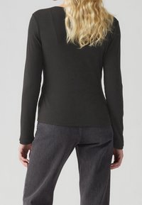 Svart långärmad topp i ribbat tyg, med en uringad rygg och sidofåror, som bärs med mörkgrå byxor.