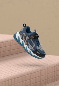 Baskets de sport bleues et grises avec une semelle épaisse, des accents texturés, un laçage et une sangle réglable, exposées sur des marches en carreaux roses.