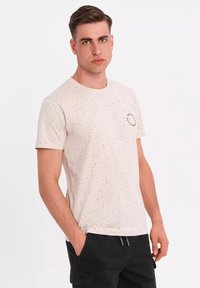Lichtbeige T-shirt met zwarte spikkels, ronde halslijn, korte mouwen en een kleine grafische print op de linkerborst. Textuurstof.