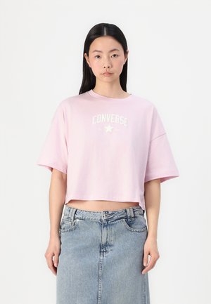 Pink cropped t-shirt made of cotton with short sleeves. Features white text design and star logo. Worn with a light blue denim skirt.