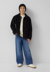 Jongen in een zwarte beanie, zwarte corduroyjas, wit shirt, wijde blauwe jeans en witte sneakers, staand tegen een lichtgrijze muur.
