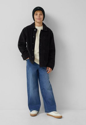 Junge, der eine schwarze Mütze, eine schwarze Cordjacke, ein weißes Shirt, weite blaue Jeans und weiße Sneaker trägt, steht vor einer hellgrauen Wand.