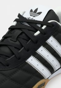 Tenisii negri Adidas cu trei dungi albe, șireturi negre și logo-ul Adidas pe limbă și talpa de cauciuc.