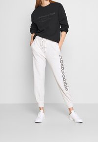 Sudadera negra con logo en relieve, combinada con pantalones de chándal gris claro que presentan texto vertical "Abercrombie". Zapatillas blancas completan el conjunto.