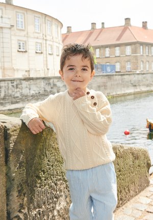 Ung dreng i en cremefarvet sweater og stripede bukser læner sig op ad et stengærd ved en kanal med historiske bygninger i baggrunden.