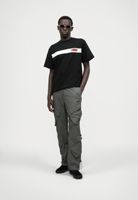 T-shirt preto de algodão com risca horizontal branca e logótipo vermelho, combinado com calças cargo cinzentas de corte largo, com vários bolsos e fechos de zíper.