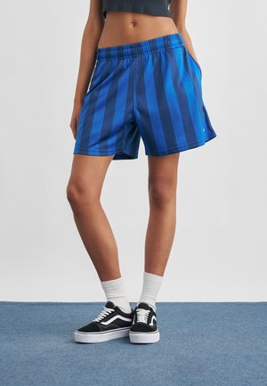 Persoon draagt blauw-zwarte gestreepte mesh korte broek, witte crew-sokken en zwarte lage sneakers, staand op een blauw tapijtvloer.