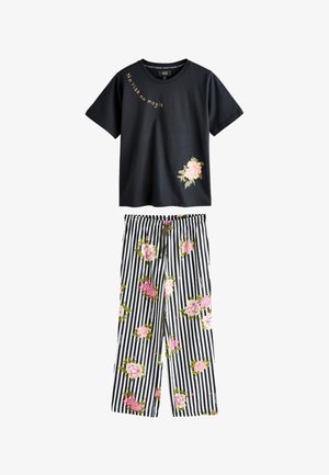 Frau steht barfuß mit verschränkten Armen, trägt ein schwarzes Hemd und gestreifte Hosen, die mit pinken Blumen verziert sind, vor einer dunklen vertäfelten Wand.