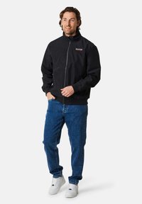 Giacca nera con zip e colletto alto, con un piccolo logo ricamato rosso sul petto. Indossata con jeans in denim blu e sneaker bianche.