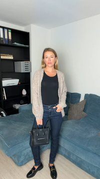 Hall hall kardigan musta pluusi peal, tumedate teksade ja mustade mokassiinidega. Kandmas väikest musta käekotti, seistes sinisel diivanil.