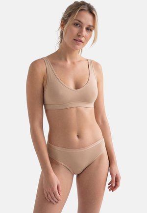 Donna che indossa un reggiseno beige senza cuciture e biancheria intima coordinata, in piedi contro uno sfondo grigio chiaro uniforme con postura rilassata.
