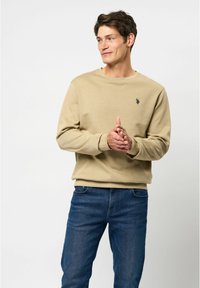 Beige sweatshirt met ronde hals en lange mouwen, voorzien van een klein geborduurd logo op de borst, gecombineerd met donkerblauwe jeans.