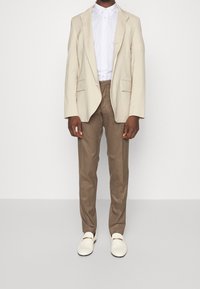 Blazer beige, camisa blanca de cuello, pantalones marrones y zapatos tipo slip-on en color blanco roto. El blazer tiene dos bolsillos frontales y corte ajustado. Texturas suaves.