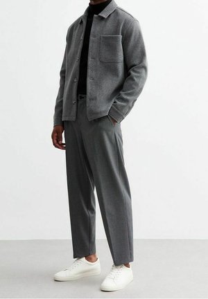 Homme portant une veste chemise grise à boutons, un col roulé sombre, un pantalon gris et des baskets blanches, debout devant un fond blanc uni.