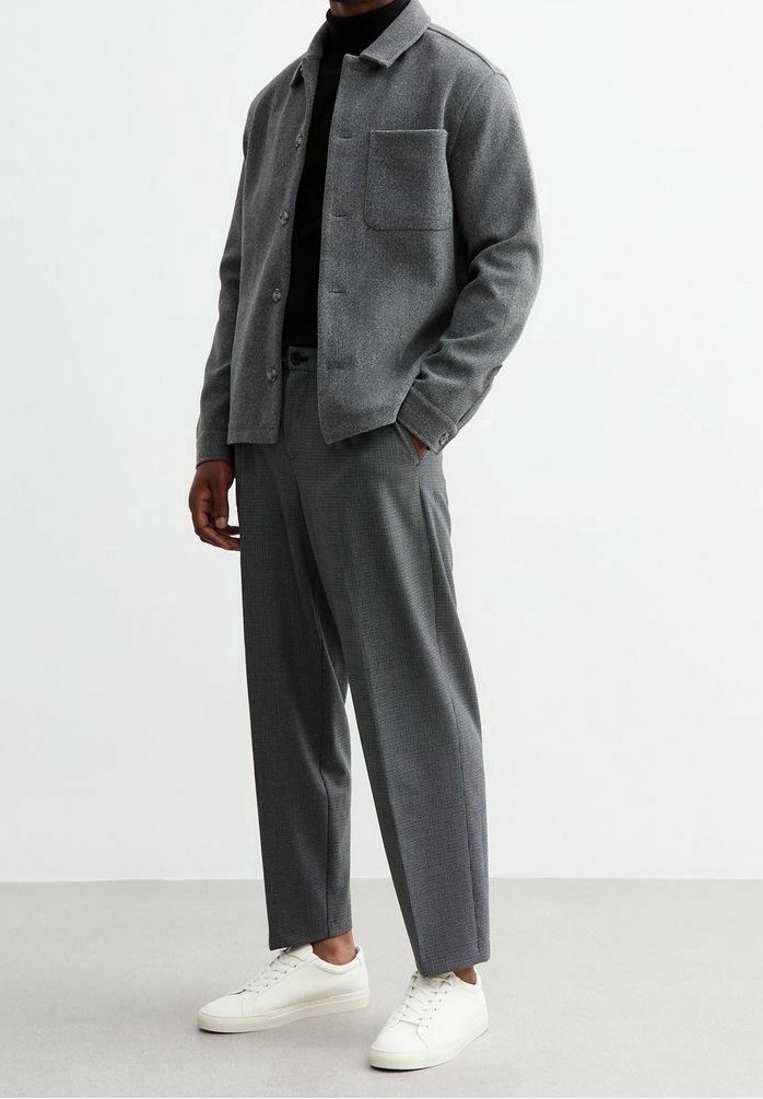 Homme portant une veste chemise grise à boutons, un col roulé sombre, un pantalon gris et des baskets blanches, debout devant un fond blanc uni.