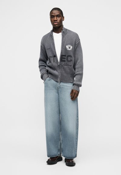 Άνδρας που φορά γκρι ριγωτό ζιπ-άπ ζακέτα με κείμενο, λευκό πουκάμισο, φαρδιά ανοιχτό μπλε τζιν και σκούρα παπούτσια, στέκεται μπροστά σε απλό φόντο.