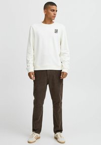 Cream sweatshirt with round neck and small graphic on front, paired with dark brown corduroy pants and white sneakers with beige accents.
