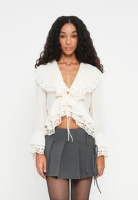 Witte gerimpelde blouse met kantafwerking en een diepe V-hals, gelaagd ontwerp en striksluiting. Gecombineerd met een grijze geplooide minirokken.