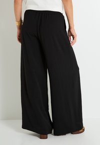 Pantalones de pierna ancha negros con una tela texturizada, con una cinturilla elástica y bolsillos laterales, llevados con zapatos de suela de madera marrones.