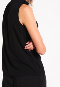 Black sleeveless top with a high neck and ruffled detailing, featuring a smooth texture and a relaxed fit. Minimalist design with no visible patterns.