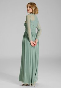 Nainen, jolla on lyhyet vaaleat hiukset ja pitkä puoliläpinä näkyvä salviavihreä mekko, seisoo kädet ristissä selän takana.