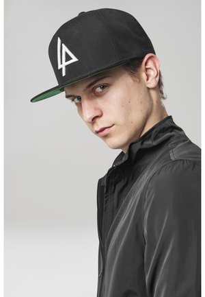 Schwarze Kappe mit grünem Unterteil, mit einem weißen Logo. Model trägt eine schwarze Jacke mit glatter Textur und Stehkragen.