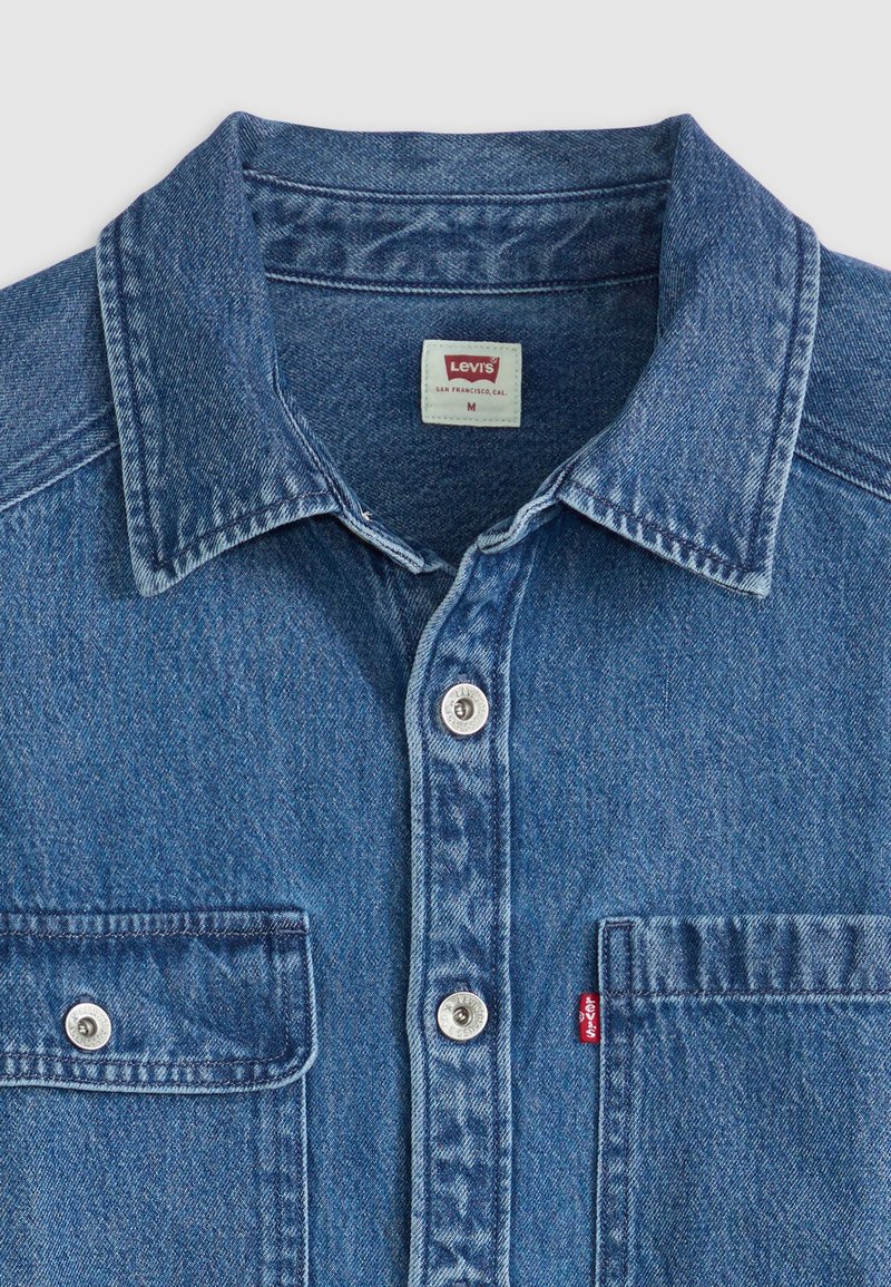 Blauwe denim shirt met een button-down kraag, twee borstzakken, zilveren drukknoopjes en een rode Levi's-label op de linkse zak.