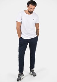 Vit kortärmad t-shirt med en liten svart triangulär logotyp, tillsammans med marinblå joggers. Grå sportskor med vita detaljer.