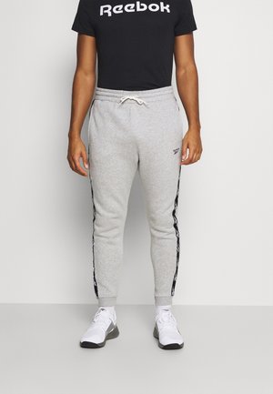 Homme portant un jogging gris avec des bandes latérales noires, des baskets blanches et un t-shirt noir Reebok, debout devant un mur uni.