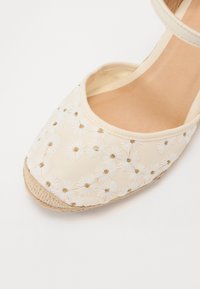 Espadrille beige con bordado floral en blanco y dorado, con parte superior de tela suave, punta redonda y suela trenzada de yute.