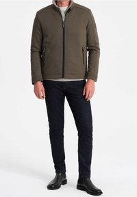 Olivgrön zip-up-jacka med texturerad design, sidofickor och hög krage, kombinerad med mörka jeans och svarta ankelboots.