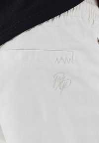 Tejido blanco con una textura suave presenta un bolsillo con un patrón de zigzag bordado y las iniciales estilizadas "RP" en hilo claro.