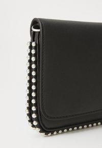 Portefeuille en cuir noir avec fermeture à rabat, présentant une rangée de clous métalliques sur un bord, texture lisse et forme structurée.