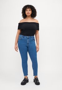 Young woman with curly hair wearing an off-shoulder black top, blue high-waisted jeans, and black studded loafers standing against white background.