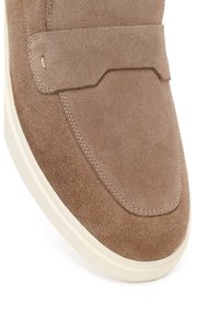 Mocasín de ante marrón claro con suela de goma blanca y detalles cosidos a lo largo de la puntera y la correa superior.