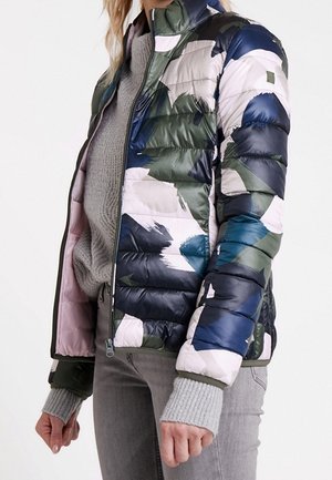 Person wearing a multicolored camo-patterned puffer jacket over a gray knit sweater and light gray jeans, standing with arms relaxed.