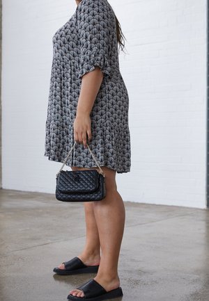 Kobieta w szarej sukience w wzory i czarnych sandałach na płaskim obcasie, trzymająca czarną pikowaną torebkę na złotym łańcuszku, stojąca w pomieszczeniu przy białej ścianie.